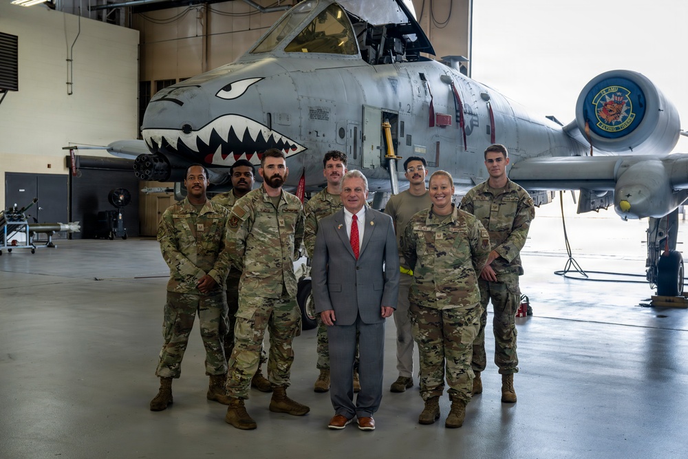 A group of seven military personnel in OCP (Operational Camouflage Pattern) uniforms pose with one civilian in a gray suit and red tie inside an aircraft hangar. They are standing in front of an A-10 Thunderbolt II (Warthog) attack aircraft featuring a distinctive shark mouth nose art. A unit patch/emblem is visible on the aircraft's tail section.