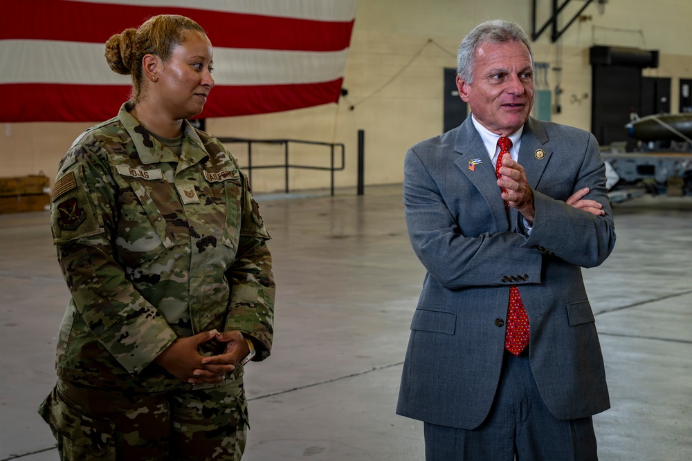 Two individuals stand inside what appears to be a military hangar with a large American flag visible in the background. A female service member in U.S. Air Force OCP (Operational Camouflage Pattern) uniform stands alongside a civilian man in a gray suit with a red tie, who appears to be speaking or gesturing. The setting suggests an official visit or briefing at a military installation.