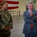 Two individuals stand inside what appears to be a military hangar with a large American flag visible in the background. A female service member in U.S. Air Force OCP (Operational Camouflage Pattern) uniform stands alongside a civilian man in a gray suit with a red tie, who appears to be speaking or gesturing. The setting suggests an official visit or briefing at a military installation.