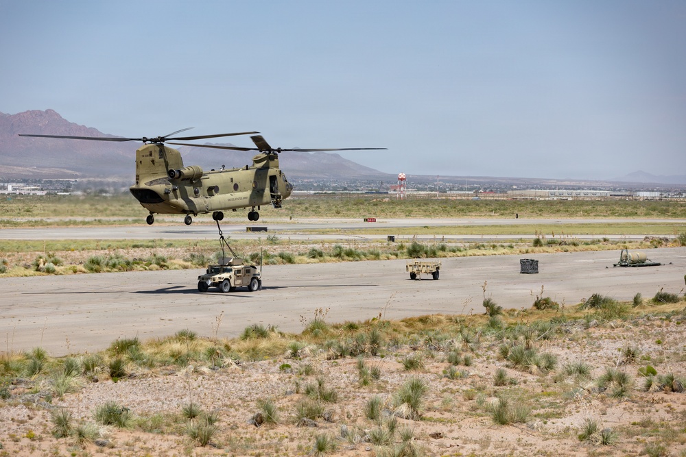 A U.S. Army CH-47 Chinook tandem-rotor helicopter is performing a sling-load operation, lifting a HMMWV (Humvee) military vehicle via a cargo hook and rigging straps on what appears to be a desert airfield or training area. A second Humvee and additional equipment are visible on the tarmac nearby. The arid, mountainous desert landscape in the background suggests a southwestern U.S. location, likely a military installation.