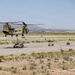 A U.S. Army CH-47 Chinook tandem-rotor helicopter is performing a sling-load operation, lifting a HMMWV (Humvee) military vehicle via a cargo hook and rigging straps on what appears to be a desert airfield or training area. A second Humvee and additional equipment are visible on the tarmac nearby. The arid, mountainous desert landscape in the background suggests a southwestern U.S. location, likely a military installation.
