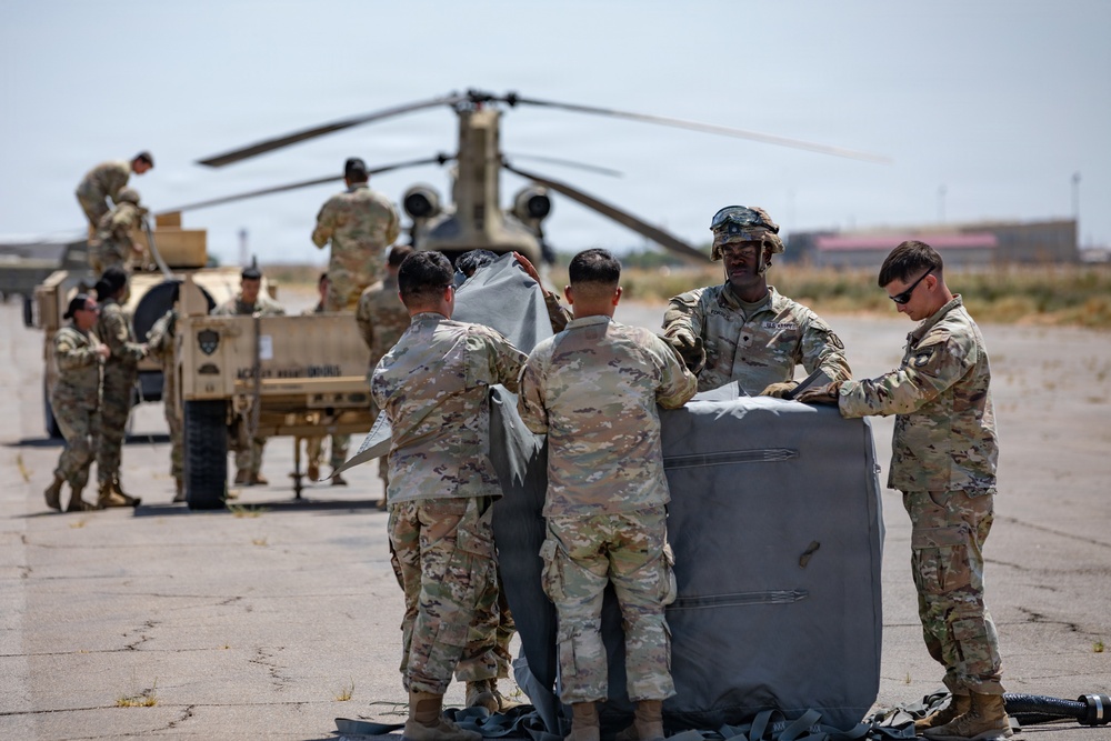 A group of U.S. Army soldiers in OCP (Operational Camouflage Pattern) uniforms are working on a tarmac or airfield, handling what appears to be a large gray equipment bundle or cargo bag. In the background, a CH-47 Chinook tandem-rotor helicopter is parked, and additional soldiers are working around a military HMMWV (Humvee) vehicle. The setting appears to be a hot, arid environment consistent with a southwestern U.S. military installation.