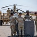 A group of U.S. Army soldiers in OCP (Operational Camouflage Pattern) uniforms are working on a tarmac or airfield, handling what appears to be a large gray equipment bundle or cargo bag. In the background, a CH-47 Chinook tandem-rotor helicopter is parked, and additional soldiers are working around a military HMMWV (Humvee) vehicle. The setting appears to be a hot, arid environment consistent with a southwestern U.S. military installation.