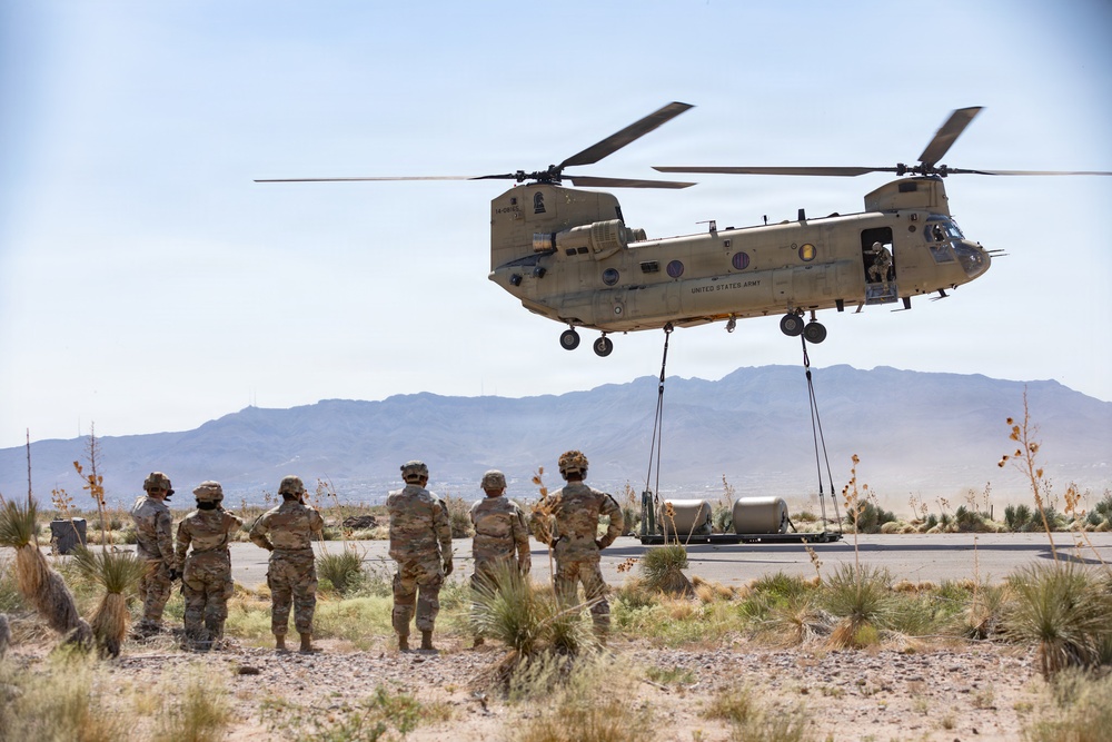 A United States Army CH-47 Chinook helicopter hovers low over a desert terrain while performing a sling-load operation, lifting military equipment secured by cables beneath the aircraft. Several soldiers in camouflage uniforms stand on the ground observing the operation. The scene appears to be a training or operational exercise in an arid, mountainous environment.