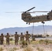 A United States Army CH-47 Chinook helicopter hovers low over a desert terrain while performing a sling-load operation, lifting military equipment secured by cables beneath the aircraft. Several soldiers in camouflage uniforms stand on the ground observing the operation. The scene appears to be a training or operational exercise in an arid, mountainous environment.