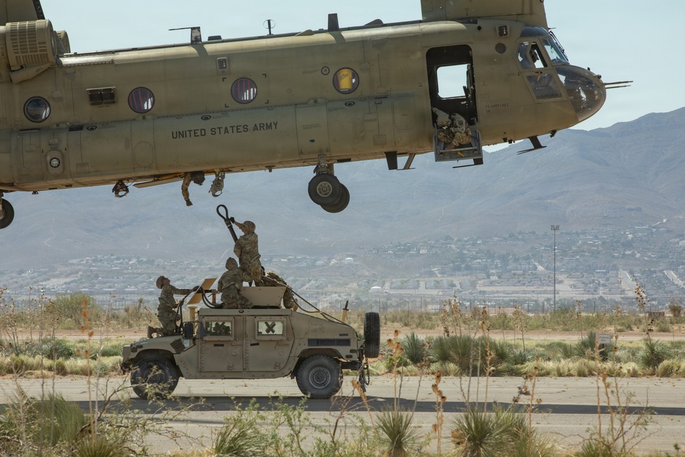 A United States Army CH-47 Chinook helicopter hovers low over a military Humvee (HMMWV) while soldiers on top of the vehicle work to attach a sling load hook. The operation appears to be a sling load or external lift training exercise taking place on a desert airfield. Mountains and a populated area are visible in the background, suggesting a location consistent with a Southwestern U.S. military installation.
