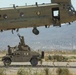 A United States Army CH-47 Chinook helicopter hovers low over a military Humvee (HMMWV) while soldiers on top of the vehicle work to attach a sling load hook. The operation appears to be a sling load or external lift training exercise taking place on a desert airfield. Mountains and a populated area are visible in the background, suggesting a location consistent with a Southwestern U.S. military installation.