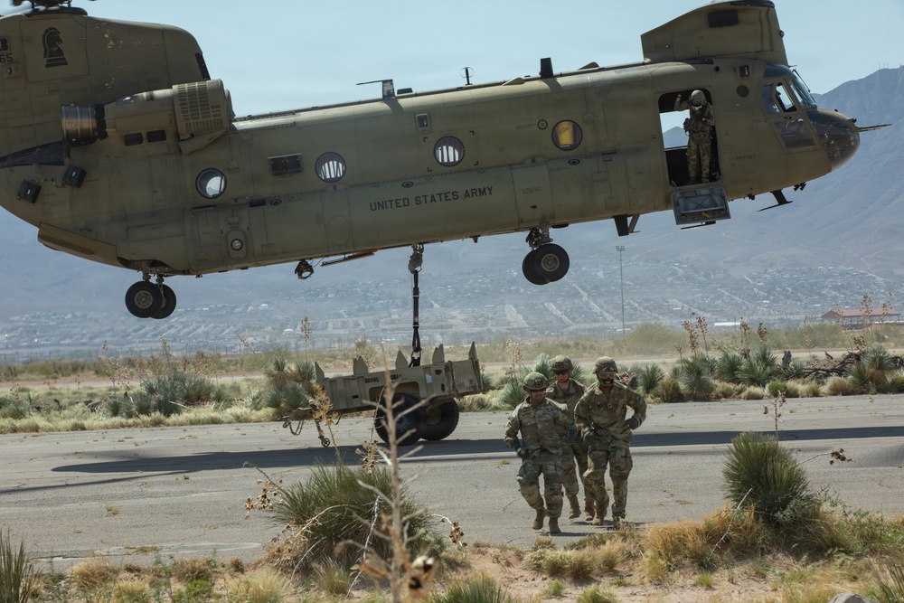 A United States Army CH-47 Chinook helicopter hovers low over a desert terrain while conducting a sling-load operation, with military equipment suspended beneath it. Three soldiers in Army Combat Uniforms (ACUs) are moving away from the helicopter on the ground below. The setting appears to be a military installation in an arid, mountainous region, likely in the American Southwest.
