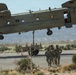 A United States Army CH-47 Chinook helicopter hovers low over a desert terrain while conducting a sling-load operation, with military equipment suspended beneath it. Three soldiers in Army Combat Uniforms (ACUs) are moving away from the helicopter on the ground below. The setting appears to be a military installation in an arid, mountainous region, likely in the American Southwest.
