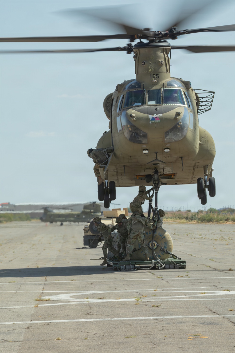 A U.S. Army CH-47 Chinook helicopter (tail number 165) hovers low over a tarmac while ground personnel attach a sling load beneath the aircraft. Several soldiers in full combat gear crouch beneath the helicopter to connect rigging to the cargo hook. At least one additional Chinook is visible in the background, suggesting a multi-aircraft sling-load training or operational exercise.