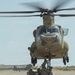 A U.S. Army CH-47 Chinook helicopter (tail number 165) hovers low over a tarmac while ground personnel attach a sling load beneath the aircraft. Several soldiers in full combat gear crouch beneath the helicopter to connect rigging to the cargo hook. At least one additional Chinook is visible in the background, suggesting a multi-aircraft sling-load training or operational exercise.