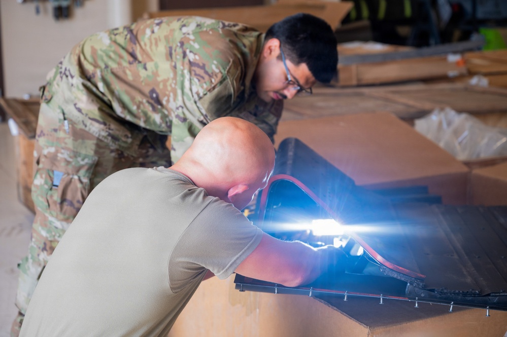 Two individuals are working together on what appears to be equipment assembly or inspection on a flat surface. One person is wearing military OCP (Operational Camouflage Pattern) uniform, while the other is in a plain olive/tan t-shirt. A bright light source is visible near the work area, suggesting use of a flashlight or work light for close inspection.
