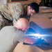Two individuals are working together on what appears to be equipment assembly or inspection on a flat surface. One person is wearing military OCP (Operational Camouflage Pattern) uniform, while the other is in a plain olive/tan t-shirt. A bright light source is visible near the work area, suggesting use of a flashlight or work light for close inspection.