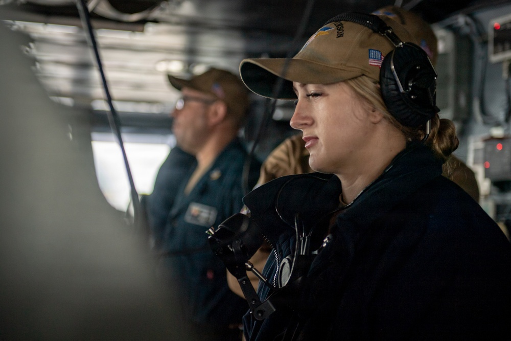 Two military personnel are stationed at what appears to be a ship's bridge or combat information center. The foreground figure is a female sailor wearing a tan cap with a US flag patch, headset/communications equipment, and dark uniform. A male sailor in a dark blue uniform is visible in the background, also wearing a cap.
