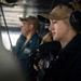Two military personnel are stationed at what appears to be a ship's bridge or combat information center. The foreground figure is a female sailor wearing a tan cap with a US flag patch, headset/communications equipment, and dark uniform. A male sailor in a dark blue uniform is visible in the background, also wearing a cap.