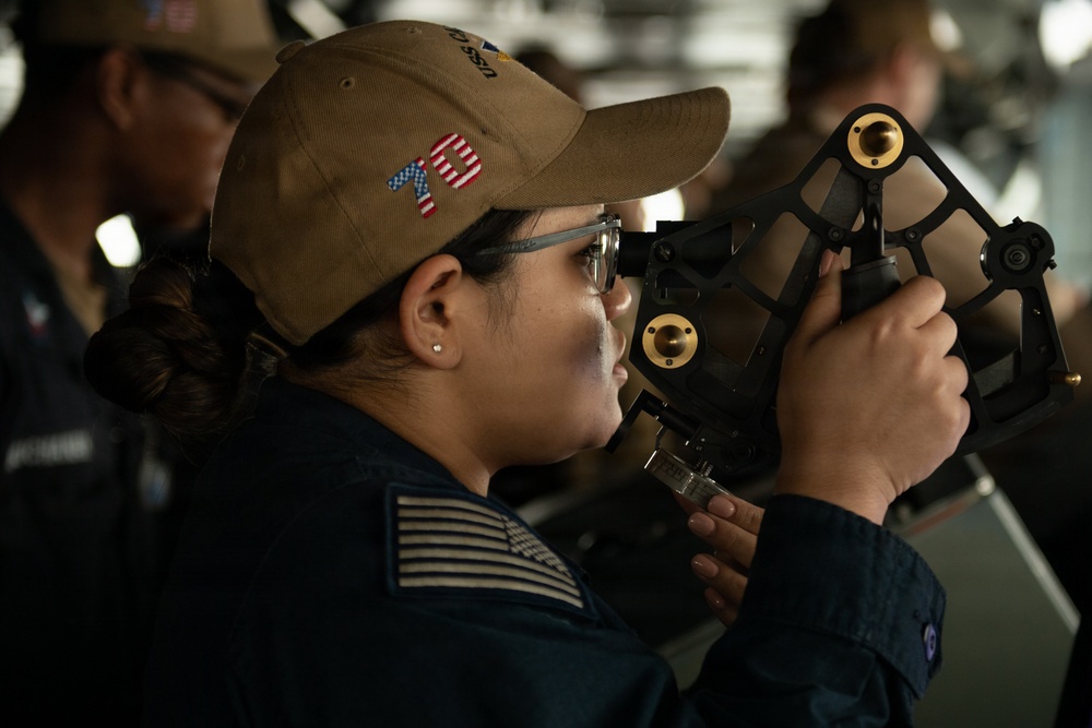 A female U.S. Navy sailor is using a sextant, a navigational instrument used to measure angles between celestial objects and the horizon. She is wearing a tan/khaki ball cap with the number '75' in American flag styling and a dark navy uniform with a U.S. flag patch on the sleeve. The setting appears to be the bridge or navigation area of a naval vessel, with other personnel visible in the background.