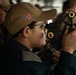 A female U.S. Navy sailor is using a sextant, a navigational instrument used to measure angles between celestial objects and the horizon. She is wearing a tan/khaki ball cap with the number '75' in American flag styling and a dark navy uniform with a U.S. flag patch on the sleeve. The setting appears to be the bridge or navigation area of a naval vessel, with other personnel visible in the background.