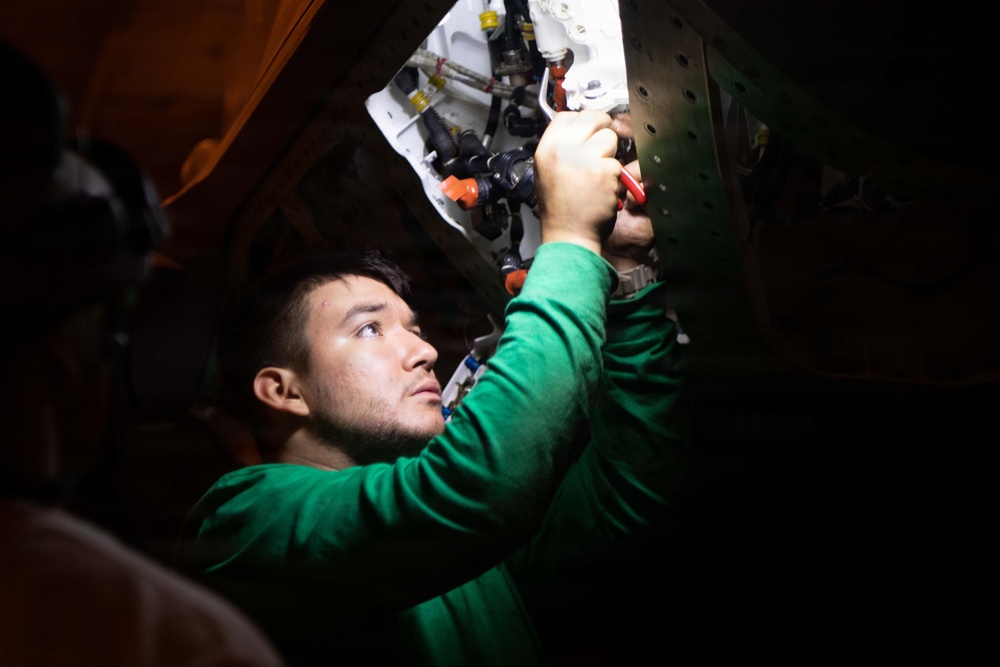A young man wearing a green long-sleeve shirt is working overhead on mechanical or plumbing components in a confined, dimly lit space. He is using a red-handled tool, likely pliers or a wrench, to work on a system of pipes, valves, and fittings mounted on a white panel. The scene suggests maintenance or repair work on a vehicle, vessel, or industrial equipment.