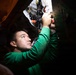 A young man wearing a green long-sleeve shirt is working overhead on mechanical or plumbing components in a confined, dimly lit space. He is using a red-handled tool, likely pliers or a wrench, to work on a system of pipes, valves, and fittings mounted on a white panel. The scene suggests maintenance or repair work on a vehicle, vessel, or industrial equipment.