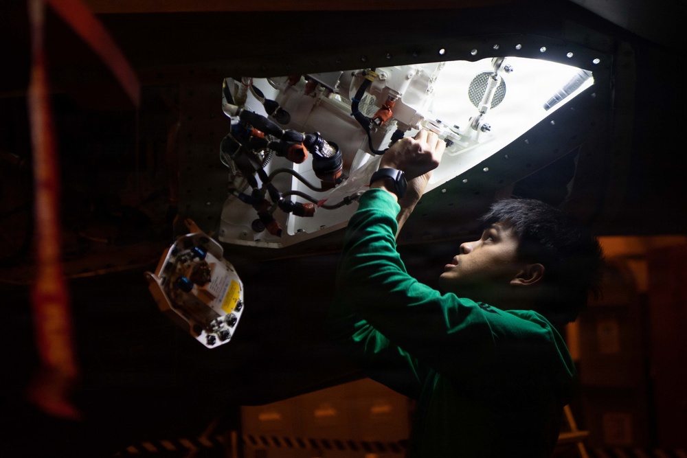 A young technician in a green long-sleeve shirt is working inside an access panel or compartment of what appears to be an aircraft or large vehicle. He is reaching upward into the illuminated interior cavity, which contains visible wiring, hoses, connectors, and mechanical components. The scene is photographed in a dark environment with the interior compartment providing the primary light source.