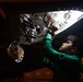 A young technician in a green long-sleeve shirt is working inside an access panel or compartment of what appears to be an aircraft or large vehicle. He is reaching upward into the illuminated interior cavity, which contains visible wiring, hoses, connectors, and mechanical components. The scene is photographed in a dark environment with the interior compartment providing the primary light source.