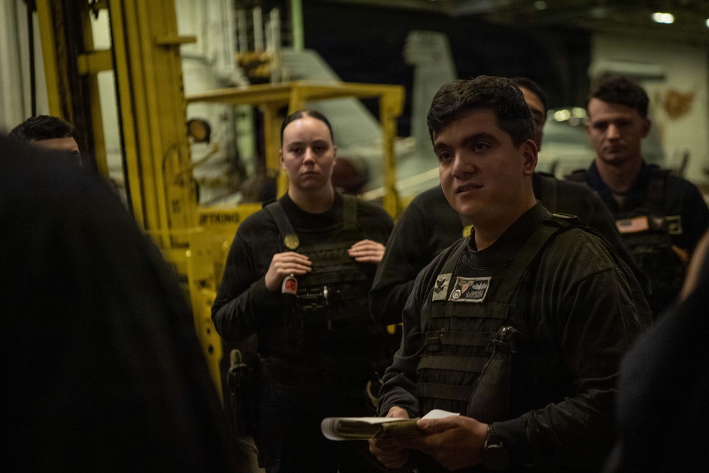 A group of military personnel in dark tactical gear and body armor are gathered in what appears to be a ship's hangar bay, likely during a briefing or meeting. The foreground figure is holding a notepad and appears to be speaking or listening attentively. Yellow industrial equipment and aircraft are visible in the background, consistent with a naval vessel environment.