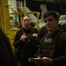 A group of military personnel in dark tactical gear and body armor are gathered in what appears to be a ship's hangar bay, likely during a briefing or meeting. The foreground figure is holding a notepad and appears to be speaking or listening attentively. Yellow industrial equipment and aircraft are visible in the background, consistent with a naval vessel environment.