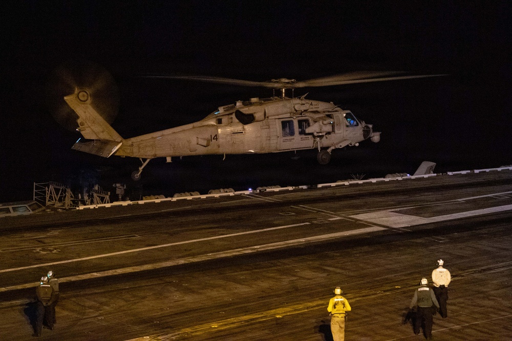 A military helicopter, marked with the number '14', is hovering or landing on the flight deck of a naval vessel at night. Several deck crew members in different colored jerseys are visible on the darkened flight deck. The scene depicts a nighttime flight operations evolution aboard what appears to be a U.S. Navy aircraft carrier or amphibious assault ship.