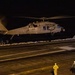 A military helicopter, marked with the number '14', is hovering or landing on the flight deck of a naval vessel at night. Several deck crew members in different colored jerseys are visible on the darkened flight deck. The scene depicts a nighttime flight operations evolution aboard what appears to be a U.S. Navy aircraft carrier or amphibious assault ship.