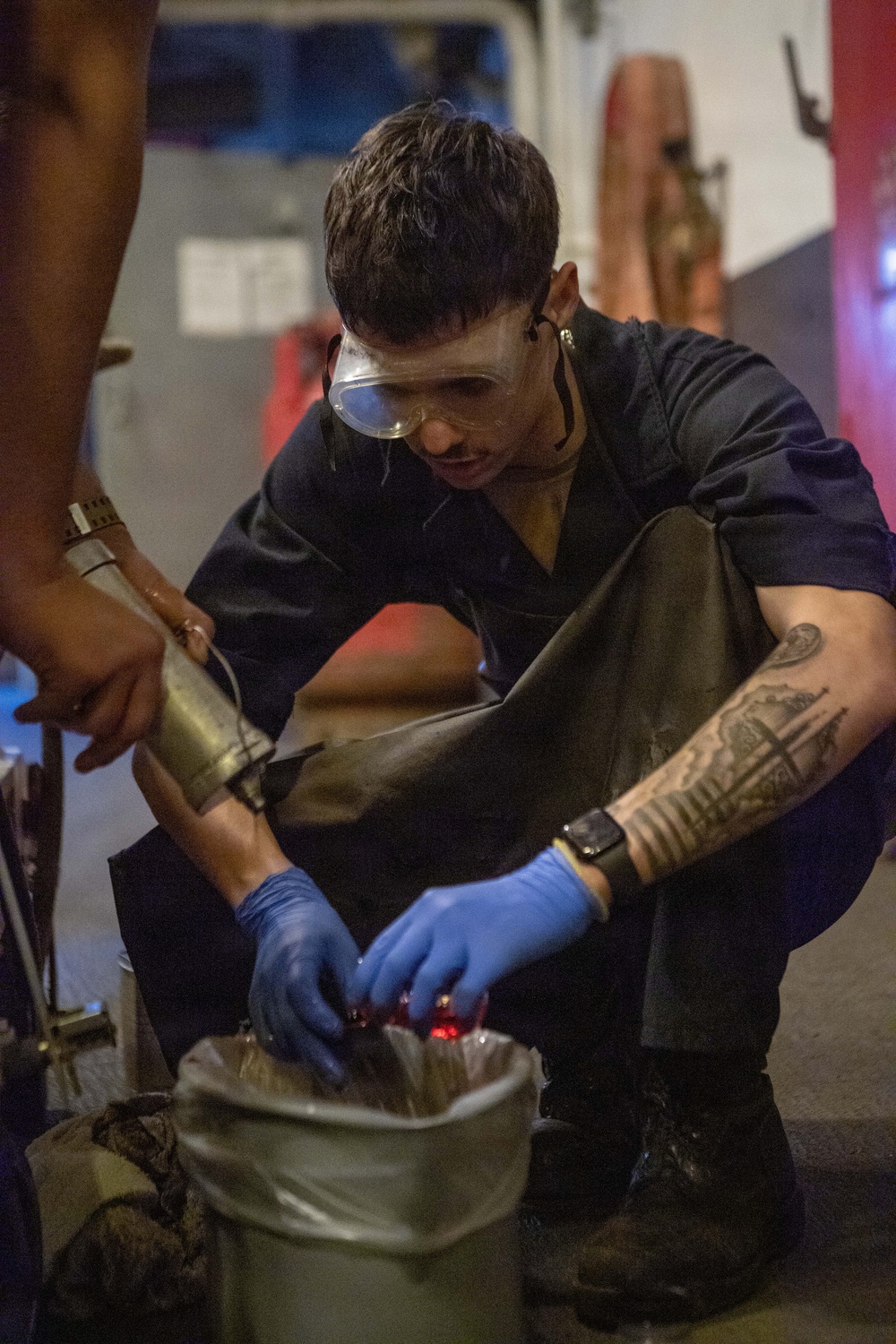 A young male technician or military/naval personnel is crouched down working with equipment in what appears to be an industrial or shipboard environment. He is wearing protective safety goggles, blue nitrile gloves, and dark work clothing, and has visible tattoos on his right forearm. He appears to be handling or inspecting items inside a container or bag with a red light source visible.