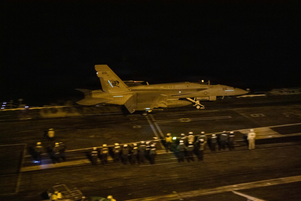 A U.S. Navy jet aircraft, marked with the number '210' and squadron designation 'VFA-83' (or similar), is photographed on an aircraft carrier flight deck at night. The aircraft appears to be either taxiing or in the process of launching or landing. Deck crew personnel in safety gear are visible along the edge of the flight deck.