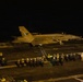 A U.S. Navy jet aircraft, marked with the number '210' and squadron designation 'VFA-83' (or similar), is photographed on an aircraft carrier flight deck at night. The aircraft appears to be either taxiing or in the process of launching or landing. Deck crew personnel in safety gear are visible along the edge of the flight deck.