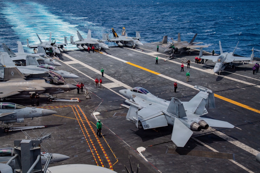An aerial view of a U.S. Navy aircraft carrier flight deck crowded with multiple fixed-wing aircraft, including F/A-18 Super Hornets and at least one F-35C stealth fighter. Deck crew in color-coded jerseys are actively working around the aircraft, indicating flight operations or aircraft handling. The carrier is underway at sea, visible by the white wake trailing behind the vessel.