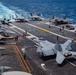 An aerial view of a U.S. Navy aircraft carrier flight deck crowded with multiple fixed-wing aircraft, including F/A-18 Super Hornets and at least one F-35C stealth fighter. Deck crew in color-coded jerseys are actively working around the aircraft, indicating flight operations or aircraft handling. The carrier is underway at sea, visible by the white wake trailing behind the vessel.