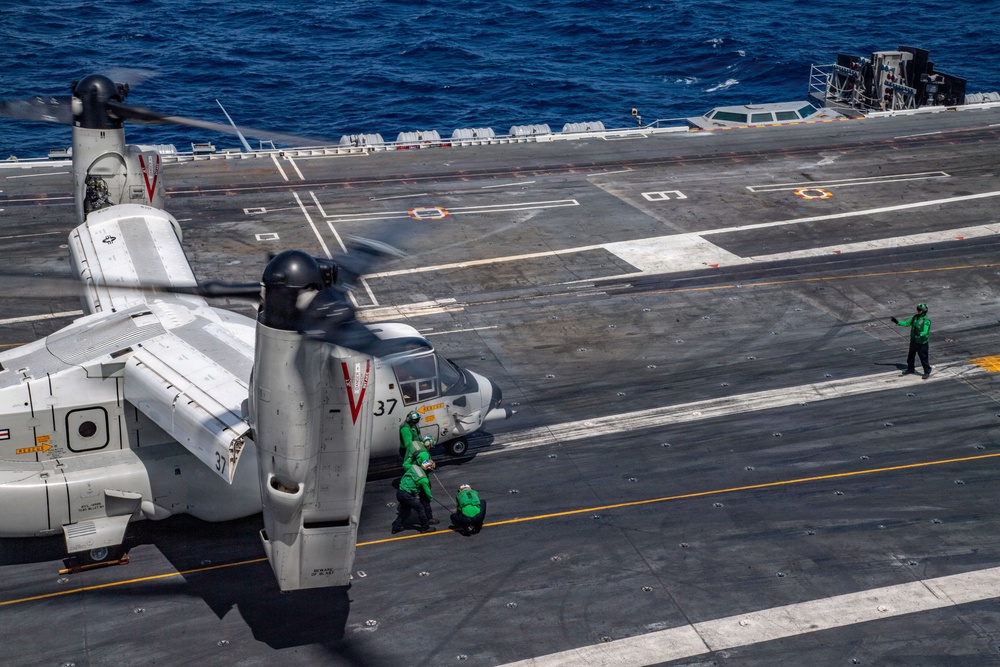 A military tiltrotor aircraft, identified as number 37 with a 'V' chevron marking consistent with a CMV-22B or MV-22B Osprey, is on the flight deck of a US Navy aircraft carrier. Ground crew members dressed in green jerseys are performing deck operations around the aircraft, which has its rotors spinning. The scene takes place at sea under clear, sunny conditions.