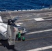 A military tiltrotor aircraft, identified as number 37 with a 'V' chevron marking consistent with a CMV-22B or MV-22B Osprey, is on the flight deck of a US Navy aircraft carrier. Ground crew members dressed in green jerseys are performing deck operations around the aircraft, which has its rotors spinning. The scene takes place at sea under clear, sunny conditions.