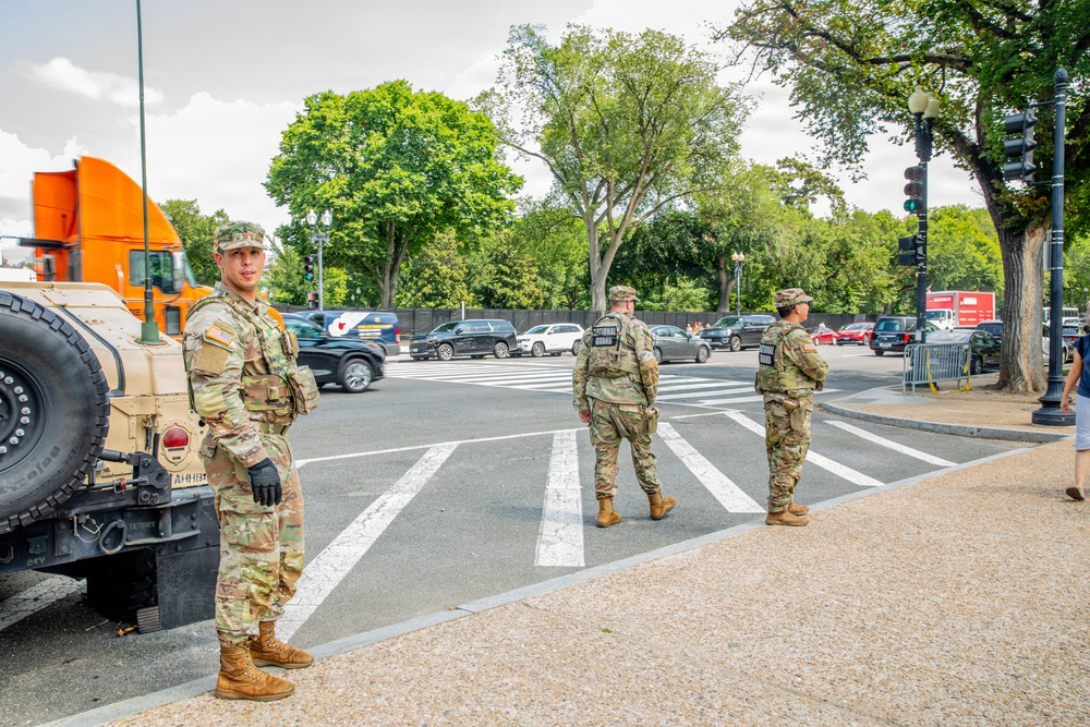 Three military personnel in MultiCam/OCP camouflage uniforms are positioned at what appears to be a street intersection in an urban area, likely Washington D.C. based on the architecture and street layout. They are standing near a tan military HMMWV (Humvee) vehicle, monitoring vehicle and pedestrian traffic at the intersection. The scene suggests a security or law enforcement support operation.