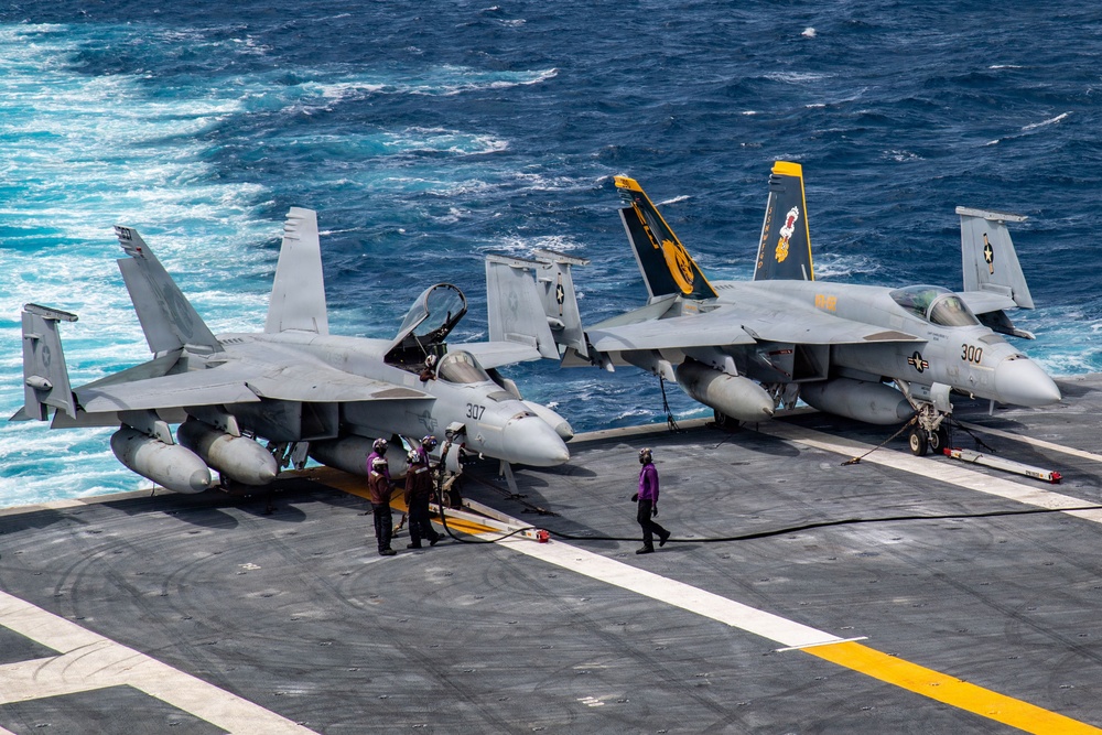 Two U.S. Navy F/A-18 Super Hornet fighter jets are parked on the flight deck of an aircraft carrier at sea. The aircraft are numbered 307 and 300, with colorful squadron markings visible on their tail fins, including yellow and orange designs. Deck crew members in colored jerseys are conducting maintenance or pre-flight operations around the aircraft.