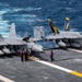 Two U.S. Navy F/A-18 Super Hornet fighter jets are parked on the flight deck of an aircraft carrier at sea. The aircraft are numbered 307 and 300, with colorful squadron markings visible on their tail fins, including yellow and orange designs. Deck crew members in colored jerseys are conducting maintenance or pre-flight operations around the aircraft.