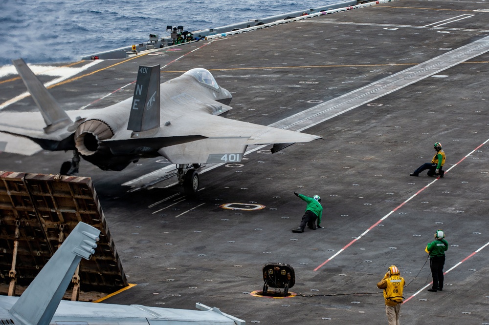 An F-35C Lightning II stealth fighter jet (bureau number 401, tail code 'NE') is positioned on the flight deck of a US Navy aircraft carrier, apparently being prepared for a catapult launch. Multiple flight deck crew members in colored jerseys are visible performing pre-launch duties. The ocean is visible in the background, confirming an at-sea operational setting.