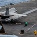 An F-35C Lightning II stealth fighter jet (bureau number 401, tail code 'NE') is positioned on the flight deck of a US Navy aircraft carrier, apparently being prepared for a catapult launch. Multiple flight deck crew members in colored jerseys are visible performing pre-launch duties. The ocean is visible in the background, confirming an at-sea operational setting.