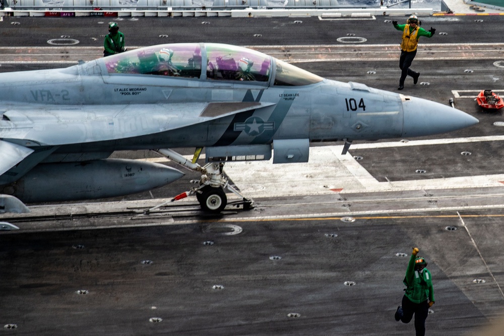 A U.S. Navy F/A-18 Super Hornet assigned to VFA-2 (Bureau Number 104) is positioned on an aircraft carrier flight deck, apparently preparing for a catapult launch. The aircraft bears pilot name markings 'LT Alec Medrano - Pool Boy' and 'LT Sean Lee - Guru' on the canopy rail. Multiple flight deck crew members in green and yellow jerseys are visible performing pre-launch duties.