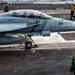 A U.S. Navy F/A-18 Super Hornet assigned to VFA-2 (Bureau Number 104) is positioned on an aircraft carrier flight deck, apparently preparing for a catapult launch. The aircraft bears pilot name markings 'LT Alec Medrano - Pool Boy' and 'LT Sean Lee - Guru' on the canopy rail. Multiple flight deck crew members in green and yellow jerseys are visible performing pre-launch duties.