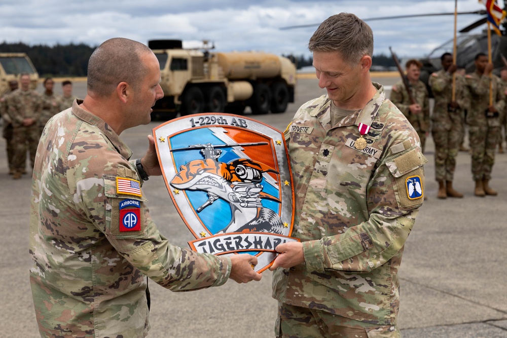 Two U.S. Army soldiers are exchanging a unit plaque/shield for the 1-229th Attack Battalion (AB) 'Tigersharks' during what appears to be a military ceremony. The soldier on the right, identified by name tape as 'Trivette,' wears Lieutenant Colonel rank and U.S. Army insignia with visible medals. The ceremony is taking place on what appears to be a military airfield with other uniformed soldiers standing in formation in the background.