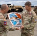 Two U.S. Army soldiers are exchanging a unit plaque/shield for the 1-229th Attack Battalion (AB) 'Tigersharks' during what appears to be a military ceremony. The soldier on the right, identified by name tape as 'Trivette,' wears Lieutenant Colonel rank and U.S. Army insignia with visible medals. The ceremony is taking place on what appears to be a military airfield with other uniformed soldiers standing in formation in the background.