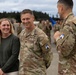 A U.S. Army military ceremony or event taking place on what appears to be a tarmac or parade ground. A male soldier in OCP (Operational Camouflage Pattern) uniform labeled 'U.S. ARMY' is smiling broadly alongside a civilian woman in a green top, while conversing with another uniformed soldier whose back is to the camera. A large formation of soldiers is visible in the background.