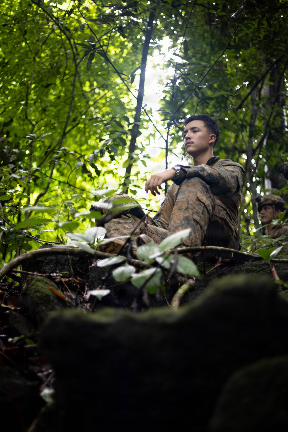 A young male soldier in military camouflage uniform is seated in a dense jungle or forest environment, appearing to be at rest or on a brief pause during a field exercise. A second soldier is partially visible in the background to the right. The low-angle shot through foreground foliage creates a dramatic, immersive composition.