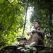 A young male soldier in military camouflage uniform is seated in a dense jungle or forest environment, appearing to be at rest or on a brief pause during a field exercise. A second soldier is partially visible in the background to the right. The low-angle shot through foreground foliage creates a dramatic, immersive composition.