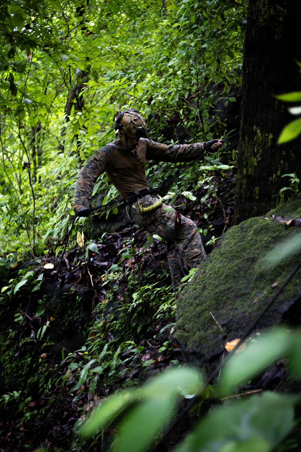 A single individual wearing military-style camouflage clothing and tactical gear is navigating through dense jungle or forest terrain. The person appears to be moving through thick vegetation, rocky ground, and heavy undergrowth. The setting suggests a military training exercise or field operation in a tropical or subtropical forest environment.