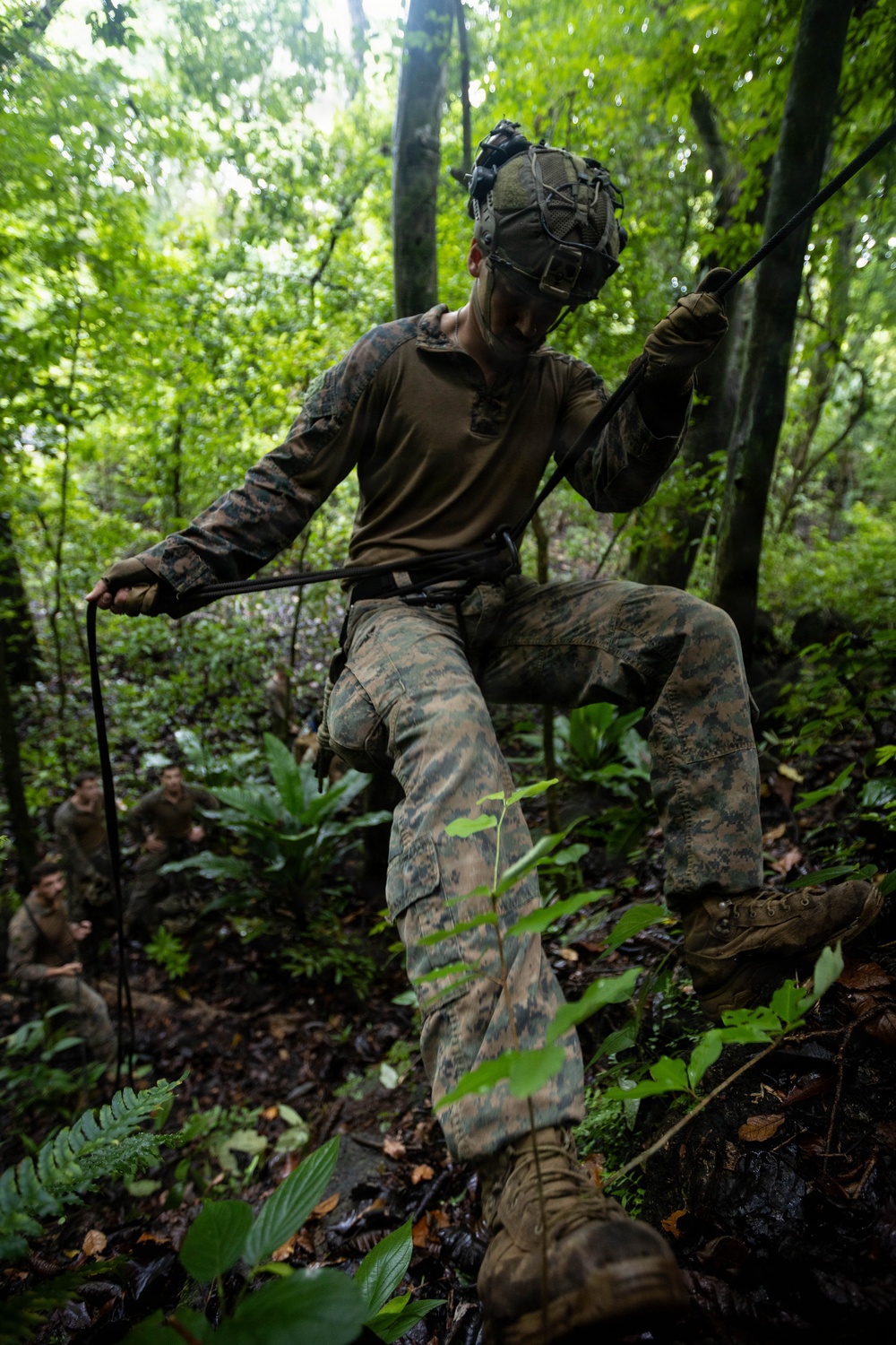 A military service member in MARPAT woodland camouflage uniform navigates a jungle terrain obstacle course using a rope or cable line. The soldier is wearing a tactical helmet with netting and gloves while traversing dense tropical forest vegetation. Additional personnel in similar uniforms are visible in the background, suggesting a group training exercise.
